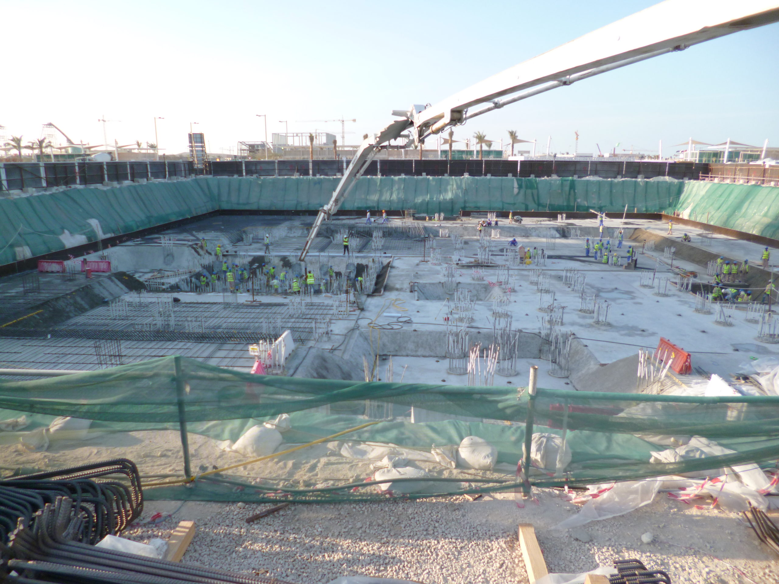 A long white crane looms over the Al Khayrien Tower worksite covered in building materials, dips in the surface, and construction workers.