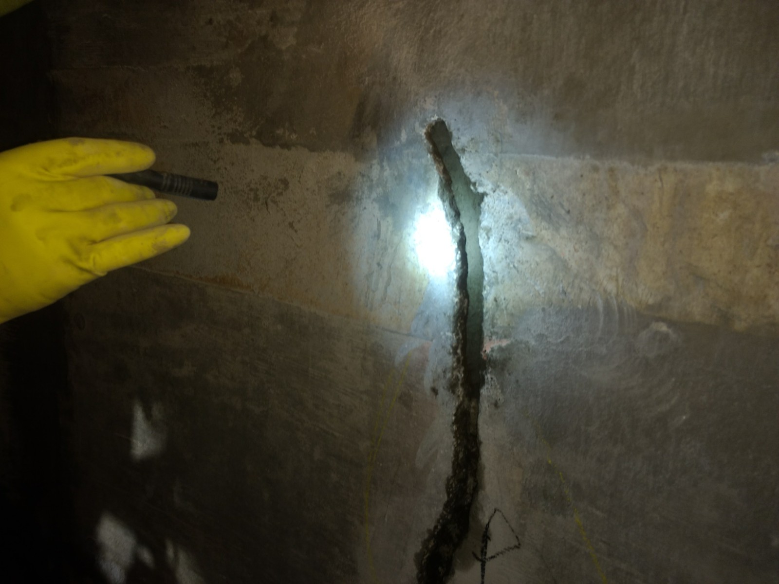 A hand in a yellow rubber glove is holding a flashlight up to a crack found in the concrete tanks of the Cameron LNG plant.