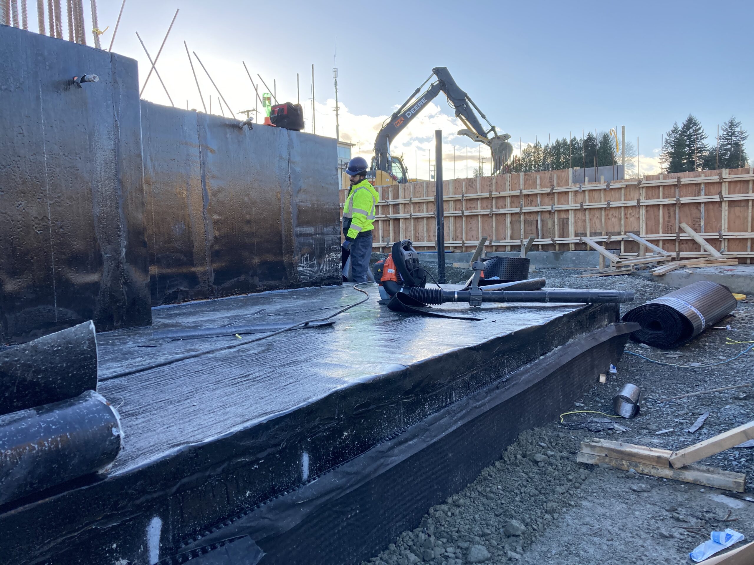 A construction worker is walking around the edges of a covered concrete structure in the middle of a worksite with a crane in the background.